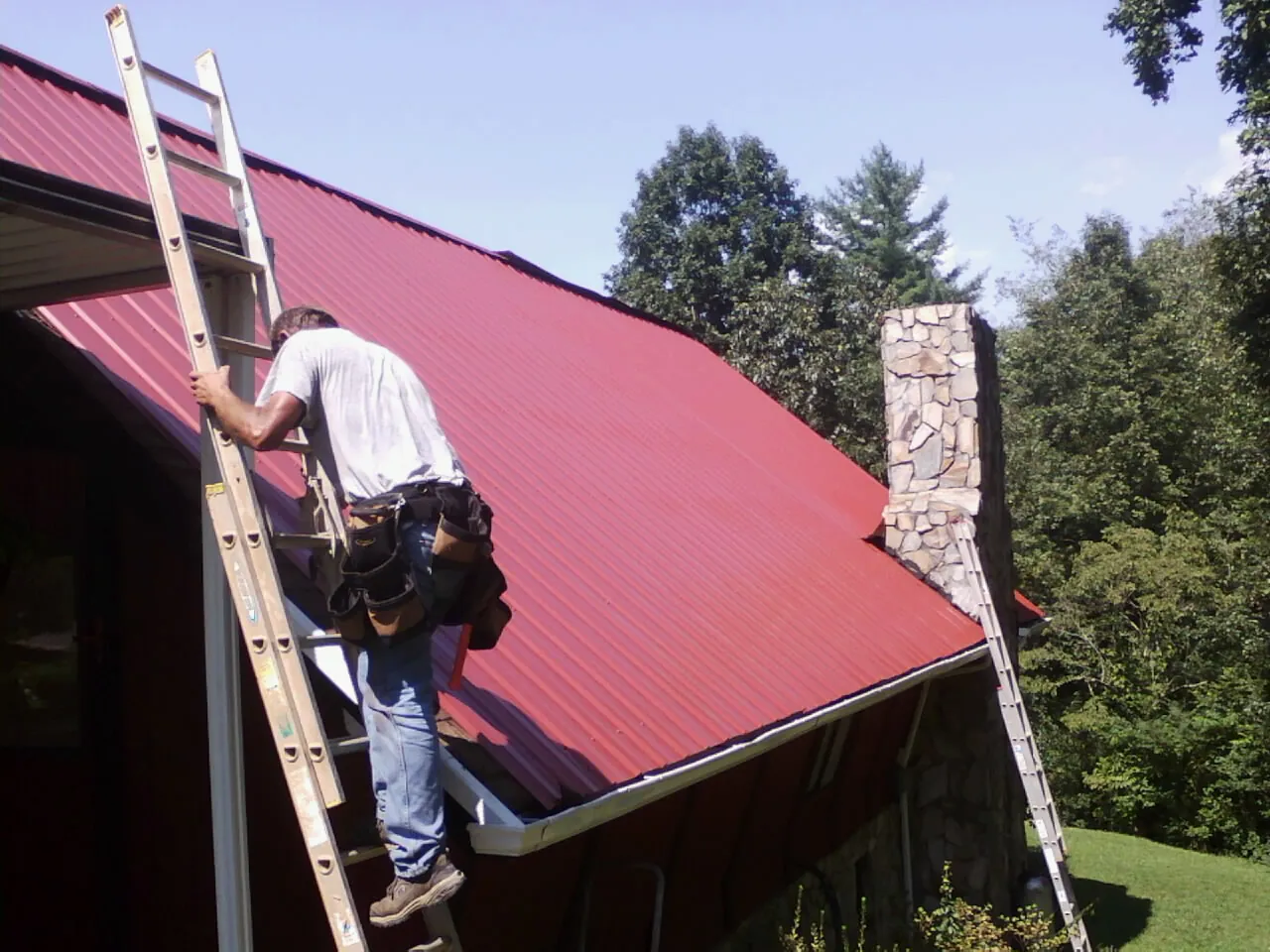 Roofer on a ladder beside a red metal roof and stone chimney — Soffit & Fascia Repair by Roofing Brothers in Linwood